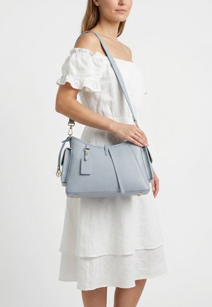 Vrouw draagt een witte off-shoulder jurk en houdt een lichtblauwe leren handtas met gouden hardware over haar schouder.
