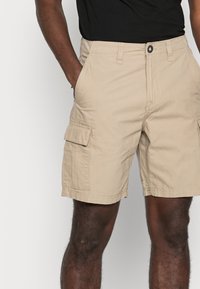 Homme portant un short cargo beige et une chemise noire, se tenant avec une main dans une poche contre un fond uni.