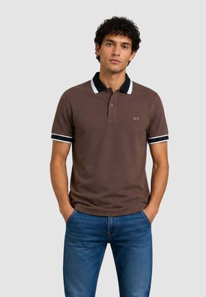 Jeune homme portant un polo marron avec un col et des bandes noires et blanches aux manches, debout les mains dans les poches d'un jean bleu.