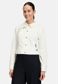 Veste en denim blanc coupée avec fermeture à boutons, poches poitrine et détails brodés noirs d'un palmier et accents de texte sur le col.