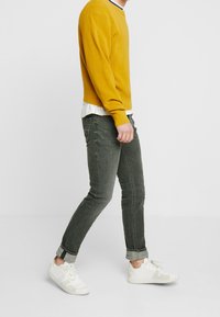 Senapsgul tröja med ribbad textur, kombinerad med mörkgröna slim-fit jeans. Vita sneakers med grå detaljer. Modell i profil.