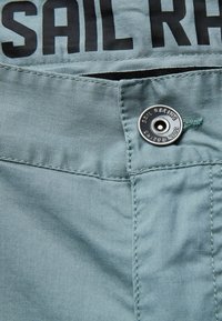 Tissu bleu clair avec une texture lisse, comportant un bouton en métal et un texte imprimé en noir "SAIL RACING". Comprend une poche zippée dissimulée.