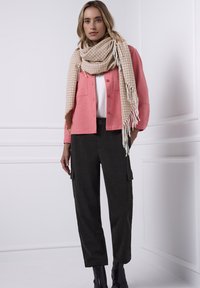 Koralowy sweter rozpinany na guziki noszony na białej bluzce, zestawiony z teksturowanymi ciemnymi spodniami typu cargo. Beżowy szalik w wzory z frędzlami dopełnia strój.