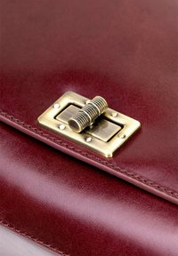 Burgundowa skórzana torba z mosiężnym zapięciem, szczegółowym przeszyciem oraz gładką, błyszczącą teksturą w prostokątnym kształcie.