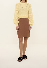 Żółty dziergany sweter z wzorzystymi rękawami, zestawiony z brązową dopasowaną spódnicą. Modelka ma na sobie czarne futrzane klapki z plecionym paskiem.