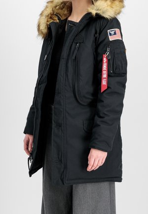 Parka noire avec capuche bordée de fourrure, fermeture éclair et boutons sur le devant, écusson avec le drapeau américain sur la manche, et étiquette rouge "Remove Before Flight" sur la poche zippée du bras.