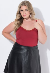 Žena s dlouhými blond vlasy, oblečená v červené krajkové tílko a černé kožené sukni, stojící proti bílému pozadí.