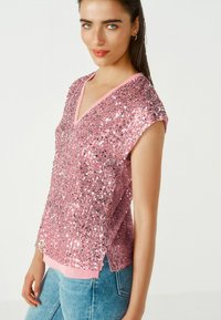 Top rose orné de sequins avec un décolleté en V et des manches courtes, présentant une base en tissu lisse et des détails de ourlet superposés.