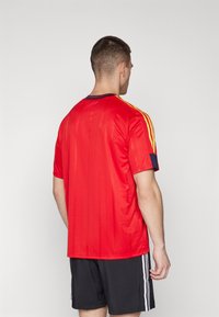 Červený sportovní dres s vertikálními pruhy, krátkými rukávy, kulatým výstřihem a žlutými akcenty na ramenou. Nosený s černými šortkami.