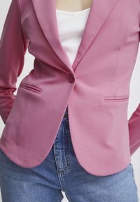 Roze blazer van gladde stof, met een enkele knoopsluiting en twee voorkruiszakken, gecombineerd met een witte geribbelde top en blauwe jeans.