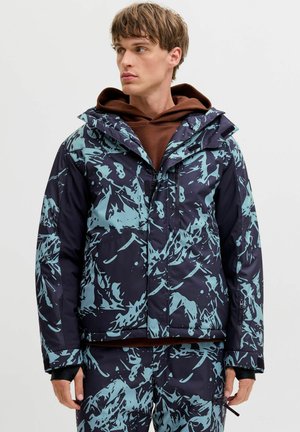 Veste de ski avec un motif abstrait bleu marine et bleu clair, dotée d'une fermeture éclair à l'avant, d'une capuche et de poignets ajustables. Le matériau semble isolé.