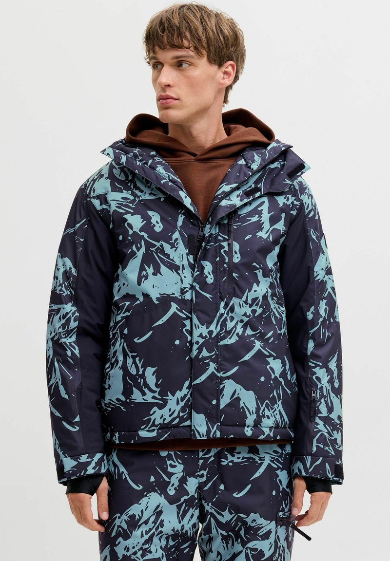 Giacca da sci con un motivo astratto blu navy e azzurro, dotata di zip frontale, cappuccio e polsini regolabili. Il materiale sembra essere isolato.