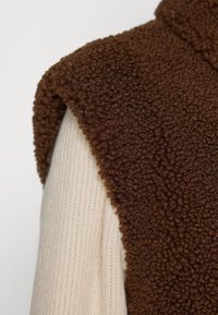 Close-up van een bruine geribbelde fleecevest gedragen over een crèmekleurige geribde, gebreide top met lange mouwen tegen een effen achtergrond.