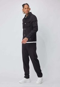 Veste en denim noir avec deux poches poitrine, assortie à un pantalon noir coordonné, et des baskets blanches ; design épuré et minimaliste.