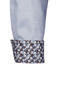 Camicia azzurra chiara con un motivo a pois, caratterizzata da un polsino decorato in tonalità di marrone, blu e un design floreale bianco.