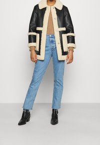 Giacca in shearling di pelle nera con accenti crema, chiusura a bottoni frontale e due tasche anteriori. Abbinata a jeans azzurri chiari e stivaletti neri.
