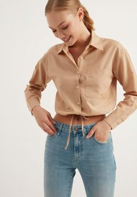 Camicia cropped di colore marrone chiaro con un piccolo taschino, annodata in vita. Abbinata a jeans a vita alta di colore blu chiaro, con una texture di denim liscia.