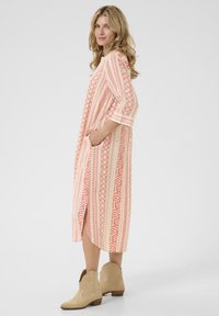 Rochie lungă, cu model în nuanțe de coral și crem. Prezintă desene geometrice, mâneci trei sferturi, buzunare laterale și o fanta. Asortată cu cizme bej.
