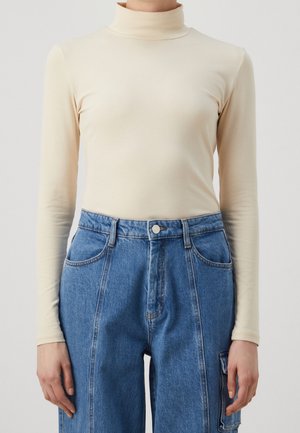Personne portant un col roulé crème ajusté et un jean denim bleu taille haute avec poches avant et latérales, mains détendues le long du corps.