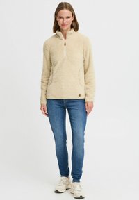 Beige fleece pullover with a high collar, zip closure, and side pockets. It has a soft, textured appearance and a subtle brand tag. Worn with blue jeans.