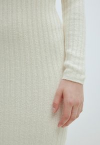Rippstrickkleid in hellem Creme, mit langen Ärmeln und taillierter Silhouette. Weicher, strukturierter Stoff mit einem subtilen vertikalen Muster.