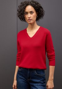 Czerwony, prążkowany sweter z dekoltem w kształcie V i długimi rękawami, zestawiony z niebieskimi dżinsami. Materiał wydaje się miękki i dopasowany.