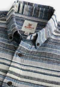 Koszula z guzikiem w paski w odcieniach niebieskiego, białego i czarnego. Wykonana z tkaniny woskowanej, z kołnierzykiem i metką z napisem "Next Supply".