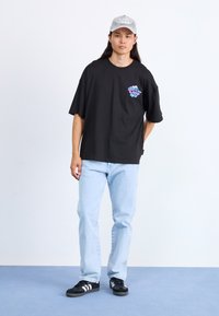 Młody mężczyzna z długimi włosami, noszący szarą czapkę, czarny T-shirt z nadrukiem, jasnoniebieskie dżinsy i czarne buty Adidas, stoi na niebieskiej podłodze.