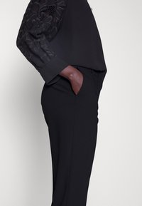 Personne portant un pantalon noir et un chemisier noir avec des manches en dentelle florale, main placée négligemment dans la poche, sur un fond uni.