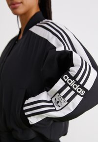 Svart bomberjakke med hvite striper langs skuldrene, Adidas-logo på ermet, og ribbede mansjetter og kant. Glatt stofftekstur.