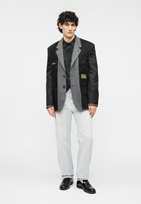 Δίχρωμο oversized σακάκι σε μαύρο και γκρι ύφασμα με λεπτές ρίγες, με κλείσιμο με κουμπιά, τσέπες και διακοσμητική λεπτομέρεια patch. Φορεμένο με ανοιχτόχρωμα τζιν.