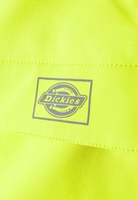 Tessuto giallo fluorescente con un logo "Dickies" grigio su un'etichetta rettangolare. Texture liscia con cuciture visibili.