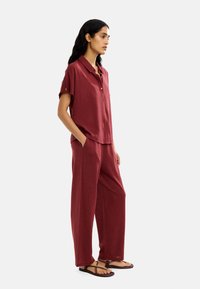 Czerwona lniana koszula na krótki rękaw z guzikami zestawiona z luźnymi, czerwonymi spodniami. Stylizacja ma swobodny charakter i została uzupełniona płaskimi sandałami.