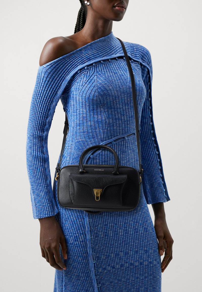 Borsa a tracolla in pelle nera con una forma strutturata, hardware dorato e due manici superiori. Indossata con un vestito in maglia blu texturizzato con design off-shoulder.