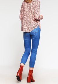 Donna che si allontana indossando una blusa floreale, jeans attillati blu e stivaletti con tacco rossi su uno sfondo chiaro e semplice.