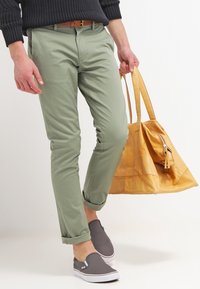 Ljust gröna chinos med slim fit och uppvikt mudd, bärs med grå slip-on skor och håller en gul läderväska.