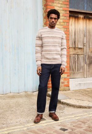 Young man in striped beige sweater, dark jeans, and brown leather shoes standing on a street with brick and wooden doors behind him.