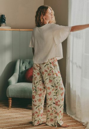 Frau in weißem, lockerem Oberteil und gemusterten Blumenhosen, steht barfuß neben einem durchsichtigen Vorhang und streckt die Hand aus; in der Nähe ein grüner Sessel und ein rundes rotes Kissen.