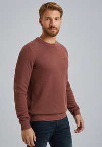 Rustfarbener Strickpullover mit Rundhalsausschnitt und gerippten Bündchen, der ein strukturiertes Muster und ein kleines Logo auf der Brust aufweist.