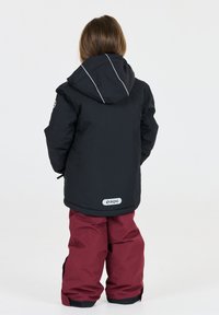 Schwarze wasserdichte Jacke mit Kapuze und reflektierenden Akzenten, kombiniert mit kastanienbraunen Hosen. Mit LOGO-Detail auf der Rückseite.