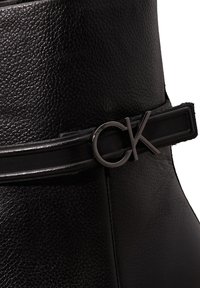 Bottine noire en cuir avec une finition texturée, dotée d'une fine ceinture noire en accent et d'un détail logo CK métallique.