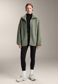 Oversized, geweven groene fleece jacket met een hoge kraag en rits, gecombineerd met zwarte leggings en lichte sneakers.