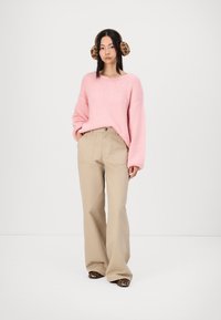 Maglione lavorato a maglia rosa chiaro con una vestibilità rilassata e una texture visibile, abbinato a pantaloni beige a gamba larga e scarpe con stampa animale.