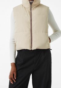 Beżowa puchowa kamizelka z wysokim kołnierzem i zapięciem na zamek z przodu, z poziomymi przeszyciami i dwiema bocznymi kieszeniami, noszona na białej koszuli z długim rękawem.