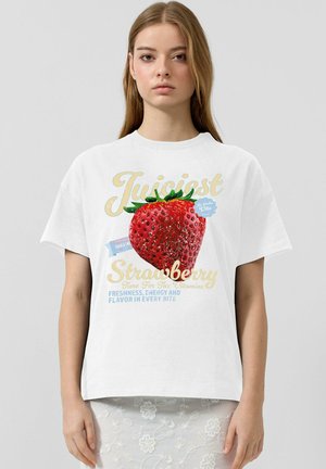 Młoda kobieta w białej koszulce z dużym czerwonym truskawkowym nadrukiem oraz napisem o świeżości i witaminach na jednolitym tle.