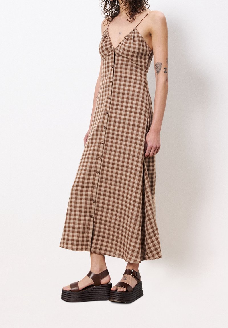 Femme portant une robe boutonnée à carreaux marron avec de fines bretelles et des sandales compensées marron, debout devant un fond blanc uni.