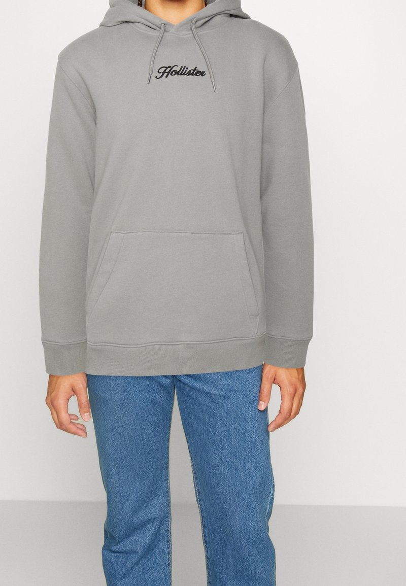 Persona con sudadera gris de Hollister con bolsillo frontal y jeans azules, de pie contra un fondo blanco liso.