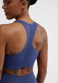 Blå idrottsbehå med racerback-design, med strukturerade paneler och perforeringar för andningsförmåga, som framhäver den passande silhuetten.