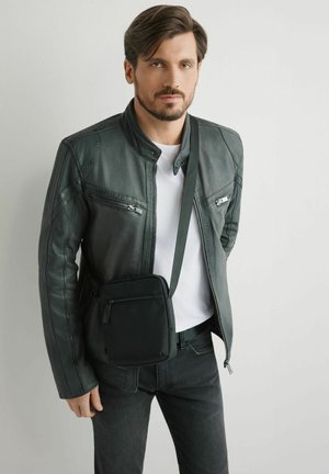 Mężczyzna o krótkich brązowych włosach i brodzie, ubrany w czarną skórzaną kurtkę, białą koszulę, czarne spodnie oraz czarną torbę na ramię na jednolitym tle.