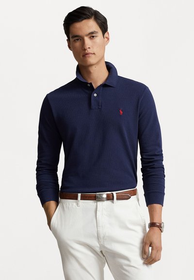 Polo à manches longues marine avec un col et un petit logo rouge. Associé à un pantalon blanc et une ceinture en cuir marron.